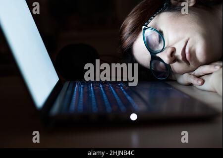 Femme fatiguée dormant sur le lieu de travail près d'un ordinateur portable. Fille a fait un boulot pour le travail à distance.Un étudiant surpassé s'est endormi sur la table.Lunettes au premier plan. Banque D'Images