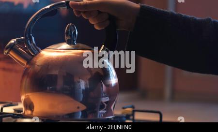 Femme plaçant la théière sur la cuisinière à gaz dans la cuisine.Gros plan de la jeune femme reflétée dans la surface polie de la bouilloire en acier la mettant sur une tourne Banque D'Images