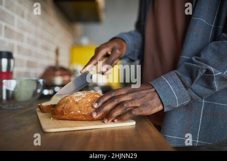 Gros plan sur un homme afro-américain méconnu qui coupe un pain frais fait maison tout en cuisinant dans la cuisine, dans un espace de travail Banque D'Images