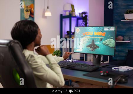 Femme joueuse excitée et réussie assise à un bureau dans un studio de jeu à la maison jouer à l'espace jeu de tir jeux vidéo gagnant tournoi en ligne.Joueur gagnant utilisant un équipement informatique RVB.Jeu d'espace cybernétique Banque D'Images