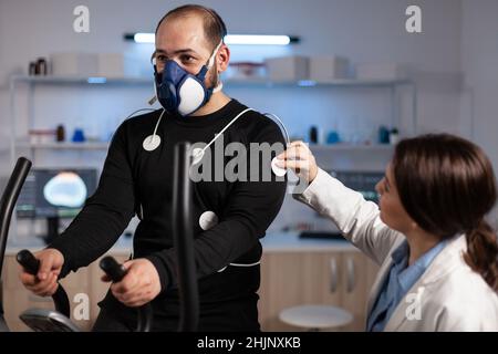 Femme spécialisée chercheuse mettant des électrodes capteur sur le sportif avec masque de surveillance des données de l'EGK dans le laboratoire médical mordern.Athlète homme en train de courir sur un entraîneur sans finale travaillant à l'endurance du corps Banque D'Images