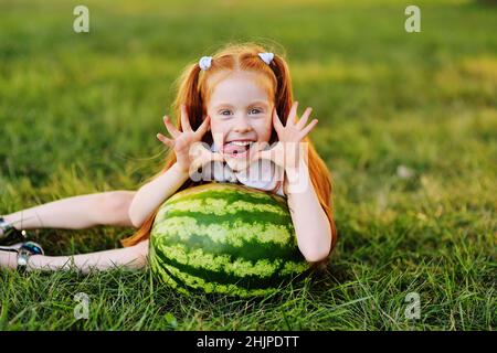 Une jeune fille à tête rouge avec des grimaces de pastèque et sourires assis sur l'herbe dans le parc Banque D'Images