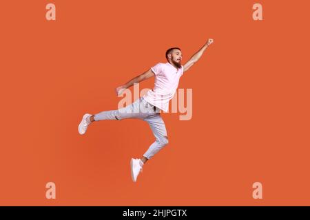 Portrait d'un homme à barbe motivé et confiant, volant dans l'air comme super-héros avec une main levée, sensation de super-puissance, portant un T-shirt rose.Studio d'intérieur isolé sur fond orange. Banque D'Images