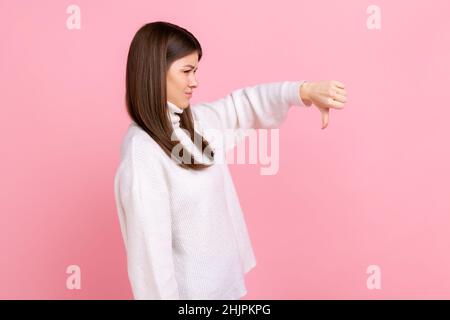 Portrait de la vue latérale de la femme bouleversée montrant le pouce vers le bas, exprimant de mauvaises émotions négatives, n'aime pas, portant blanc style décontracté pull.Studio d'intérieur isolé sur fond rose. Banque D'Images