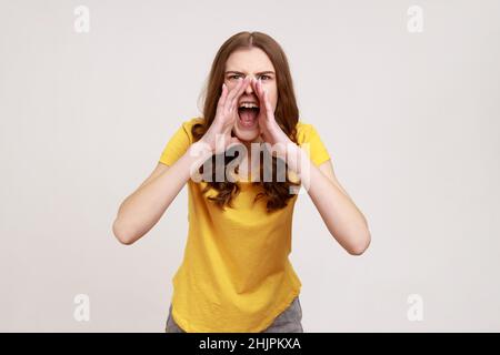 Portrait d'une jeune fille furieuse irritée avec des cheveux ondulés bruns dans un t-shirt jaune décontracté criant bruyamment, éprouvant une forte agression.Prise de vue en studio isolée sur fond gris. Banque D'Images