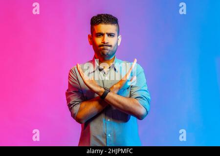 Non, absolument pas.Portrait de l'homme en chemise montrant le signe x avec les mains croisées, ce qui signifie arrêt, c'est la fin. Prise en studio en intérieur isolée sur fond coloré de lumière néon. Banque D'Images