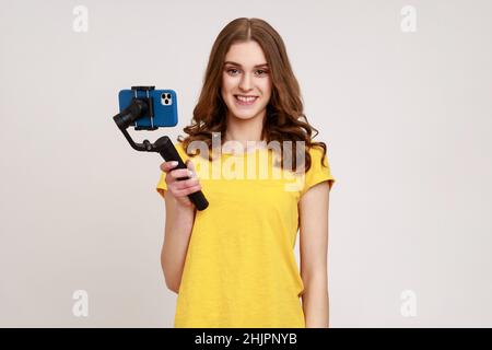 Portrait de Blogger adolescente fille en T-shirt jaune tourne vidéo pour vlog sur stabilisateur électronique, tenant Steadicam. Regardant sourire à la caméra.Prise de vue en studio isolée sur fond gris. Banque D'Images