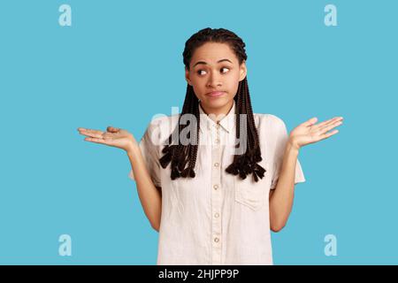 Le portrait d'une belle femme confuse avec des dreadlocks noirs se sent incertain car ne peut pas trouver la bonne façon, levant les bras, portant une chemise blanche.Studio d'intérieur isolé sur fond bleu. Banque D'Images