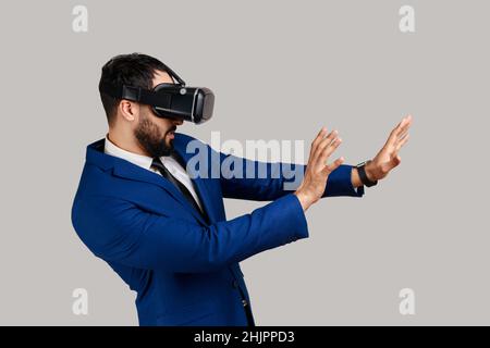 Vue latérale de l'homme barbu dans des lunettes vr, étirant les mains vers l'avant, essayant d'attraper quelque chose tout en jouant à la réalité virtuelle, peur visage choqué.Prise de vue en studio isolée sur fond gris. Banque D'Images