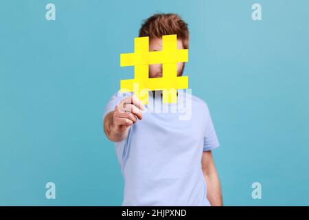 Portrait de l'homme barbu se cachant derrière le symbole jaune hashtag, couvrant le visage avec le signe de hash, le concept de blogging et les sujets viraux sur Internet.Studio d'intérieur isolé sur fond bleu. Banque D'Images
