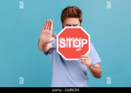 Portrait d'un homme barbu couvrant la moitié du visage avec un symbole d'arrêt octogonal, montrant un panneau de signalisation rouge et un geste d'interdiction avec la paume, avertissement de voie interdite.Studio d'intérieur isolé sur fond bleu. Banque D'Images