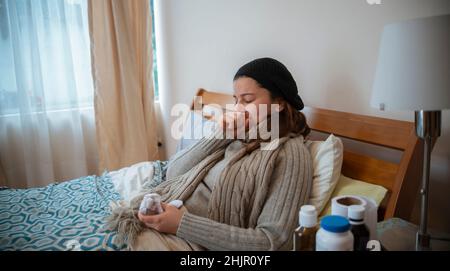 Une femme hispanique qui mentait seule dans son lit bien enveloppée, malade avec la grippe, couvrant sa bouche avec sa main tout en éternuant Banque D'Images