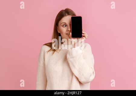 Portrait d'une drôle de femme blonde couvrant son oeil avec téléphone portable avec écran vide pour la publicité, regardant loin, portant chandail blanc.Studio d'intérieur isolé sur fond rose. Banque D'Images