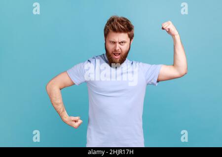 Portrait d'un homme fier et confiant, barbu, montrant le pouvoir dans ses mains, montrant des biceps, se sentant de l'énergie pour gagner le succès, posture du culturisme.Studio d'intérieur isolé sur fond bleu. Banque D'Images