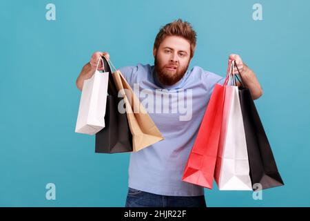 Portrait d'un homme à bout de bras barbu épuisé et à bout de bras levé avec des sacs de shopping, regardant l'appareil photo, se sent ennuyé après avoir été dans le centre commercial.Studio d'intérieur isolé sur fond bleu. Banque D'Images