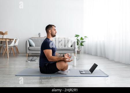 Calme millénaire caucasien gars assis, méditant sur le tapis avec ordinateur portable dans le salon intérieur, profil, espace de copie Banque D'Images