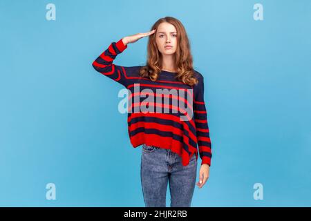 Oui, monsieur.Portrait d'une femme sérieuse portant un pull rayé de style décontracté, debout saling avec la main près de la tête, regardant obéissant et attentif à l'appareil photo.Studio d'intérieur isolé sur fond bleu Banque D'Images