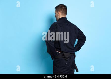 Vue arrière du jeune homme, policier portant un uniforme noir isolé sur fond bleu.Concept de travail, de frénésie, de droit et d'ordre. Banque D'Images