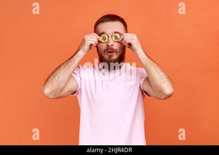 Portrait d'un homme barbu surpris couvrant les yeux avec du bitcoin doré, regardant l'appareil photo à travers la pièce de btc, portant un T-shirt rose.Studio d'intérieur isolé sur fond orange. Banque D'Images