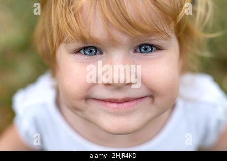 Un visage d'enfant souriant en gros plan.Belle fille heureuse avec les yeux bleus et les cheveux blonds.Un joli petit enfant souriant dans un parc, regardant directement dans l'appareil photo Banque D'Images