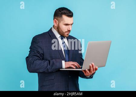 Jeune adulte concentré portant un costume de style officiel tenant un ordinateur portable dans les mains, travaillant sur l'ordinateur, regardant l'affichage avec une expression sérieuse.Studio d'intérieur isolé sur fond bleu. Banque D'Images