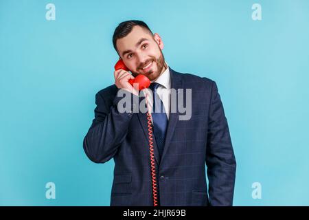 Un homme heureux avec une barbe portant un costume de style officiel parlant téléphone fixe tenant dans le combiné, regardant loin avec un sourire crasseux.Studio d'intérieur isolé sur fond bleu. Banque D'Images