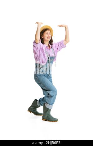 Jeune fille drôle, jardinière féminine en uniforme de travail et chapeau montre pantomime isolé sur fond blanc.Concept de travail, émotions, agronomie, éco Banque D'Images