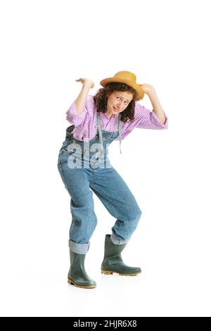 Jeune fille drôle, jardinière féminine en uniforme de travail et chapeau montre pantomime isolé sur fond blanc.Concept de travail, émotions, agronomie, éco Banque D'Images