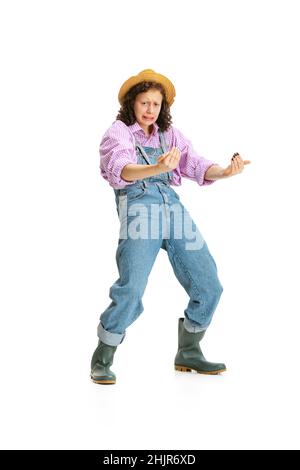 Jeune fille drôle, jardinière féminine en uniforme de travail et chapeau montre pantomime isolé sur fond blanc.Concept de travail, émotions, agronomie, éco Banque D'Images