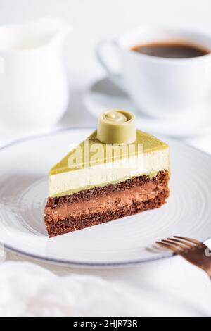 Gâteau au chocolat au pistache avec quatre couches et glaçage vert au pistache servi sur une assiette blanche sur fond blanc avec du café en flou Banque D'Images