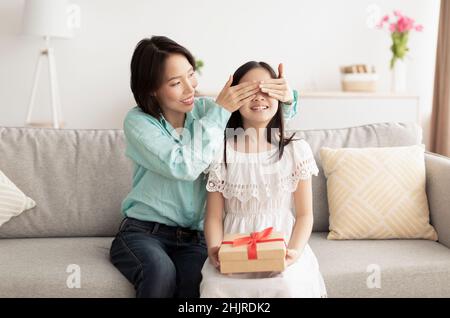 Femme asiatique mûre couvrant les yeux de sa petite-fille, faisant cadeau d'anniversaire surprise à la maison Banque D'Images