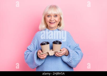 Portrait d'une jolie femme gaie aux cheveux gris tenant entre les mains deux tasses de caféine isolées sur fond rose pastel Banque D'Images