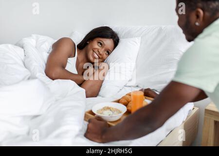Gai jeune mari noir tenir plateau avec petit déjeuner pour femme souriante sur le lit dans la chambre blanche intérieure Banque D'Images