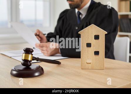 Un gavel et une maison de jouets sur une table en cour, avec un juge travaillant en arrière-plan Banque D'Images