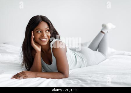 Souriante, jeune femme afro-américaine vêtue de vêtements domestiques, profitez de votre temps libre, allongez-vous au lit le week-end ou en vacances Banque D'Images