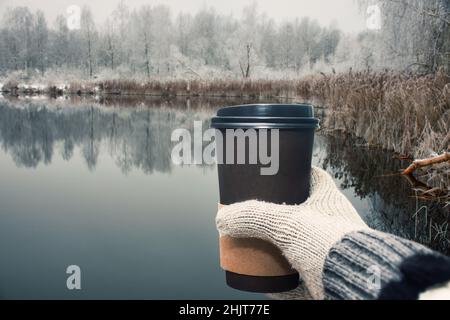 une main dans une mitaine chaude tient une tasse en papier écologique avec café sur le rivage du lac. voyage à la nature. Banque D'Images
