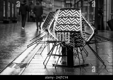 Chaises assemblées du café-terrasse dans une rue pluvieuse et piétons de derrière passer par, image de la vie de ville en noir et blanc, espace de copie, sélectionné Banque D'Images