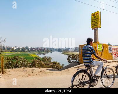 Un homme local fait un vélo sur un pont routier au-dessus d'une rivière avec vue sur les champs et les bâtiments à la périphérie de Varanasi, Uttar Pradesh, nord de l'Inde Banque D'Images