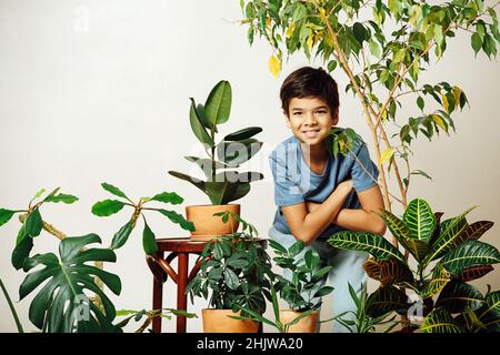 Portrat d'un garçon indien debout au milieu d'une salle pleine de plantes en pot dans des pots de céramique marron sur fond beige.Se pencher vers l'avant, se replier Banque D'Images