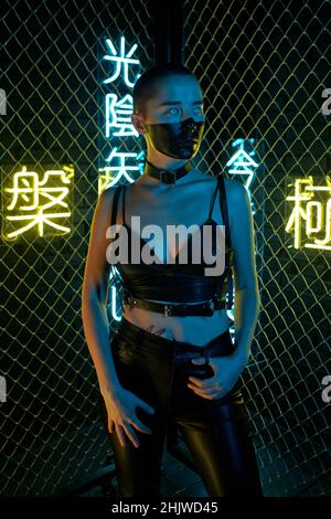 Cyber-punk fille sérieux dans un pantalon en cuir noir et une table de cuisson debout devant l'appareil photo contre les bars et les hidevers pirates Banque D'Images