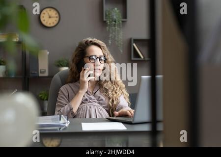 Jeune femme enceinte en robe grise décontractée parlant au téléphone portable tout en étant assise devant un ordinateur portable et en regardant l'affichage Banque D'Images