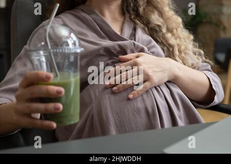 Jeune femme enceinte en robe grise gardant la main sur le ventre tout en étant assise dans un fauteuil près du bureau et en prenant un cocktail de fruits ou smoothie Banque D'Images