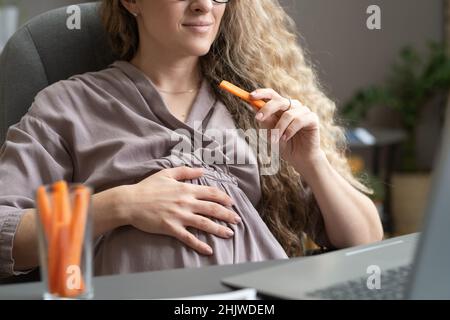 Femme d'affaires enceinte contemporaine mangeant des carottes et gardant la main sur le ventre tout en étant assise devant un ordinateur portable Banque D'Images
