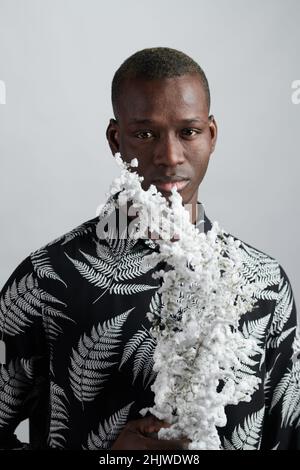 Jeune homme africain dans une chemise élégante tenant une branche couverte de fleurs blanches tout en se tenant en face de l'appareil photo dans l'isolement Banque D'Images