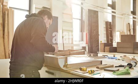 Gros plan de charpentier qualifié en uniforme noir coupant un morceau de bois dans son atelier de travail du bois.Les outils de menuisier se trouvent sur la table.Tir horizontal.Vue latérale Banque D'Images