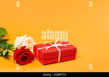 Boîte cadeau rouge avec ruban blanc et deux roses rouges et blanches sur fond orange vif.Concept cadeau de Saint-Valentin ou d'anniversaire Banque D'Images