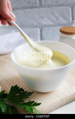 La main d'une femme tient une cuillère à café de mayonnaise sur un bol blanc avec sauce.Le concept de nutrition appropriée.Orientation verticale. Banque D'Images