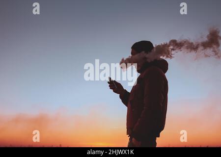 Un homme dans une veste fume une cigarette électronique à l'extérieur.Sur fond de coucher de soleil.Alternative aux cigarettes analogiques Banque D'Images