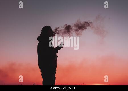 Un homme dans une veste fume une cigarette électronique à l'extérieur.Sur fond de coucher de soleil.Alternative aux cigarettes analogiques Banque D'Images