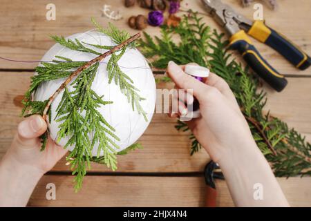 Fleuriste au travail: Femme montre comment faire boule de Noël avec sphère de styromousse et brindilles de thuja. Étape par étape, tutoriel. Banque D'Images